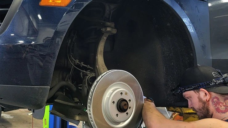 Technician wearing a headlamp working at the front wheel of a dark blue SUV with the brake rotor and caliper exposed